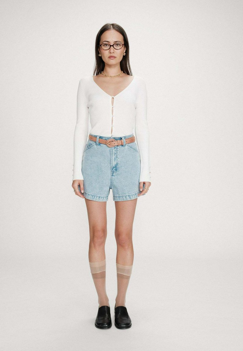 Femme debout portant un haut blanc à manches longues boutonné, un short en jean bleu clair avec une ceinture marron, des bas résille jusqu'aux genoux, des mocassins noirs, des lunettes et des bijoux.