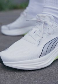 Zapatillas deportivas blancas con parte superior de malla, rayas de acento negras y una suela acolchada. Presentan cordones planos y una parte superior texturizada para mayor transpirabilidad.
