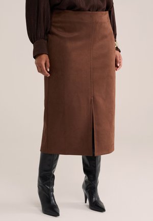 Frau trägt einen braunen knielangen Rock mit einem vorderen Schlitz, schwarze spitze High-Lederstiefel und eine dunkle langärmelige Bluse.