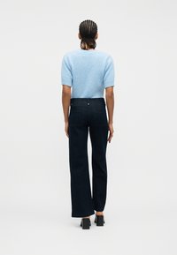 Άτομο με πλεξούδες, φορώντας ανοιχτό γαλάζιο πουλόβερ με κοντά μανίκια, σκούρο παντελόνι με ψηλή μέση και φαρδιά μπατζάκια, και μαύρα παπούτσια με τακούνι, μπροστά προς τα πίσω.