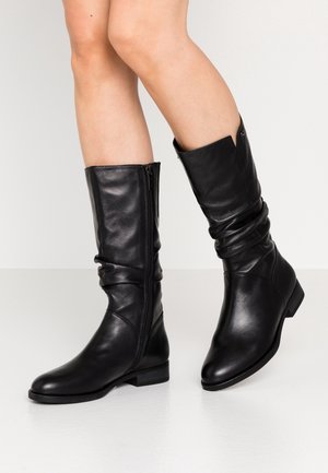 Benens met kniehoge zwarte leren laarzen met zijritsen, één voet plat op de grond en de andere iets opgetild.