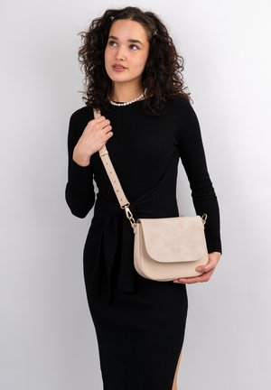 Borsa a tracolla beige con patta in suede, forma rotonda, hardware dorato e tracolla regolabile, tenuta da una persona in un vestito nero.