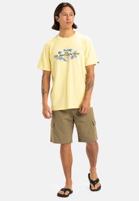 Camiseta amarilla de manga corta con diseño gráfico, combinada con pantalones cortos de cargo beige y chanclas negras, de pie sobre un fondo liso.