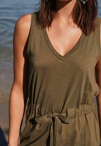 Top sans manches vert olive avec un décolleté en V, présentant une taille cintrée et un détail de nœud. Tissu doux et lisse avec une texture légère.