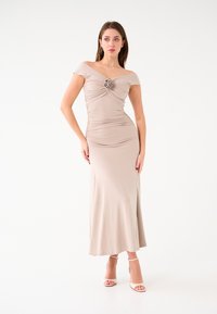 Abito beige off-shoulder con corpino arricciato e applicazione floreale. Design aderente, texture liscia e gonna lunga fino alla caviglia. Tacchi beige con cinturini.
