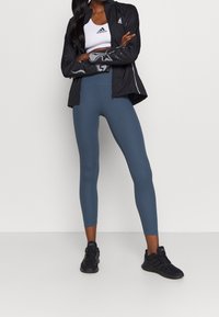 Musta kevyt takki heijastavilla yksityiskohdilla, valkoinen urheilullinen lyhyt toppi, siniset korkeavyötäröiset legginsit ja mustat lenkkarit.