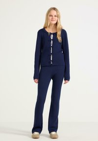 Námořnický pletený svetr s žebrovaným vzorem a předními zavazováním, spárovaný s odpovídajícími žebrovanými kalhotami. Měkká textura a přiléhavý design, vhodné pro neformální nošení.