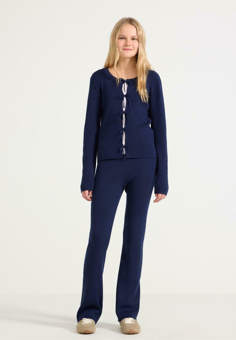 Námořnický pletený svetr s žebrovaným vzorem a předními zavazováním, spárovaný s odpovídajícími žebrovanými kalhotami. Měkká textura a přiléhavý design, vhodné pro neformální nošení.