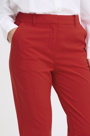 Close-up of person wearing tailored red pants with hands in pockets and a white long-sleeve shirt tucked in.