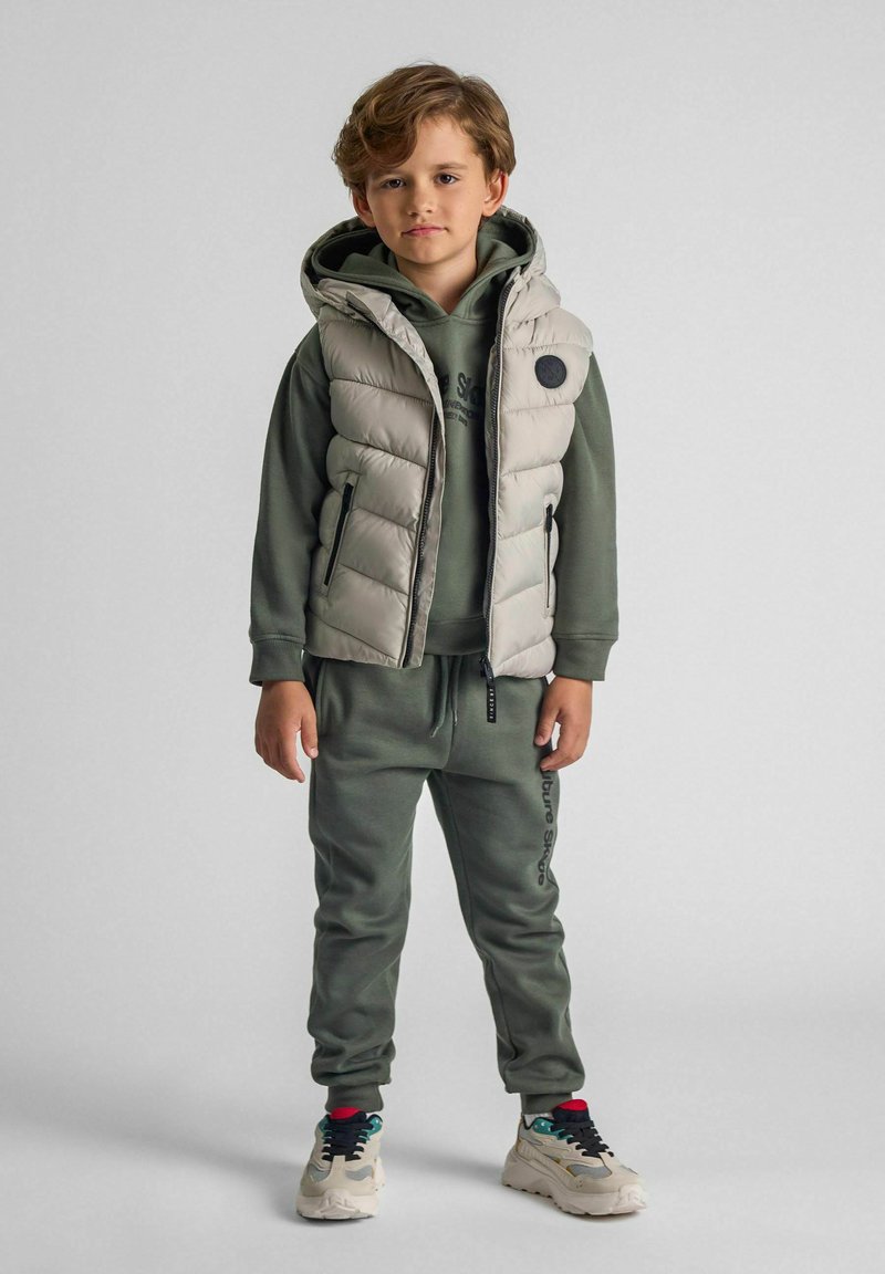 Ljusgrå vadderad väst med huva över en mörkgrön sweatshirt, parat med matchande joggers och flerfärgade sneakers.