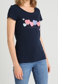 Námořnická modrá bavlněná tričko s výstřihem do srdce, na kterém je srdcový grafický motiv v růžové, modré a stříbrné barvě. Měkká textura s krátkými rukávy.