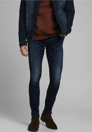 Homme portant un jean slim bleu foncé, des bottines en daim marron, un sweat-shirt marron et une veste en denim bleu foncé, une main dans la poche.
