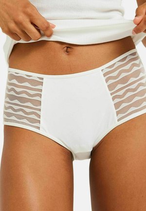 Femme soulevant un haut blanc côtelé pour révéler un sous-vêtement blanc taille haute avec des panneaux en mesh à motifs sur les hanches, vue rapprochée du torse et des cuisses.