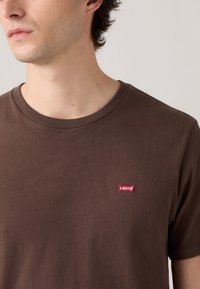 Fiatal férfi barna Levi's pólót visel, amelyen piros logócímke található a bal mellen, nyaknál és vállon rövidített.