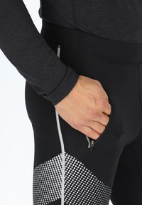 Zwarte sportleggingen met een ritszak, voorzien van een gestructureerd, gestippeld patroon in het wit langs de onderkant van het been. Rekbare stof verhoogt de bewegingsvrijheid.