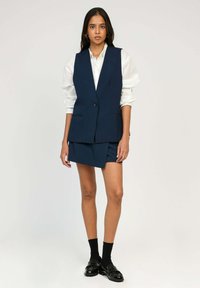 Gilet sans manches bleu marine avec boutons sur le devant, associé à une jupe asymétrique assortie. Chemisier blanc en dessous, porté avec des chaussettes noires et des chaussures.