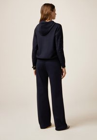 Un sweat à capuche bleu marine avec capuche et épaules ornées, associé à un pantalon bleu marine ample. La tenue présente une texture douce et une coupe décontractée.