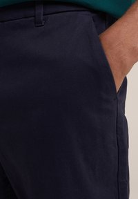 Pantaloni din bumbac bleu marin, cu o textură netedă, având un buzunar lateral și un design elegant. Prezintă o croi simplu, modern.