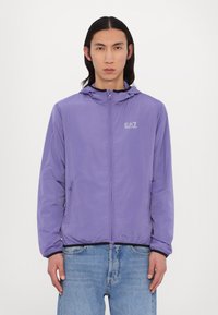 Casaco leve roxo com capuz, fecho frontal e detalhe de logótipo no peito. Apresenta punhos elásticos e bolsos laterais.