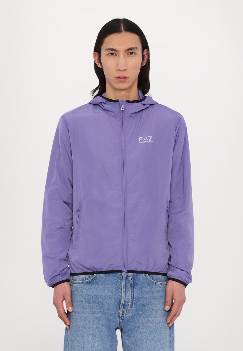 Casaco leve roxo com capuz, fecho frontal e detalhe de logótipo no peito. Apresenta punhos elásticos e bolsos laterais.