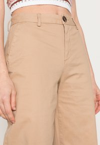 Calças largas bege feitas de um tecido leve e suave. Apresentam um fecho frontal com botão e bolsos laterais. Detalhe de costura visível.