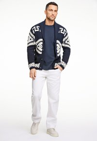 Marinblå cardigan med vitt geometriskt mönster, stickad textur, sjalkrage, knappstängning, tillsammans med marinblå skjorta och vita byxor, toppad med krämfärgade sneakers.