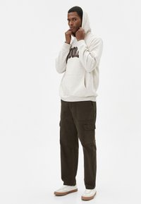 Sudadera de forro polar beige con bolsillo frontal y letras marrón oscuro, combinada con pantalones cargo en verde militar y zapatillas blancas con suelas marrón.