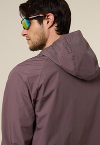 Mann trägt braune Kapuzenjacke mit "ALTAVIA"-Schriftzug auf der Kapuze, steht seitlich und trägt reflektierende Sportsonnenbrille mit orangen Akzenten.