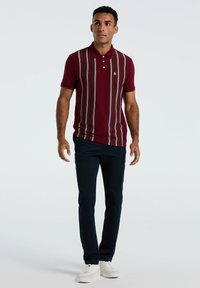 Man in een bordeauxrode kortmouw polo met verticale strepen, donkere broek en witte sneakers, staande tegen een effen achtergrond.