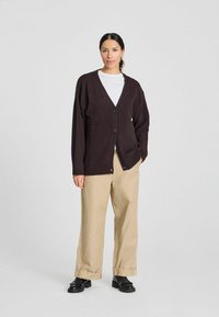 Donkerbruin gebreid cardigan met een diepe V-hals, twee knopen aan de voorkant en geribde manchetten, gecombineerd met beige wijde broekspijpen en zwarte schoenen.