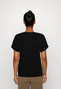 T-shirt nera in cotone con maniche corte arrotolate, scollo tondo e vestibilità comoda, mostrata di spalle, abbinata a pantaloni beige.