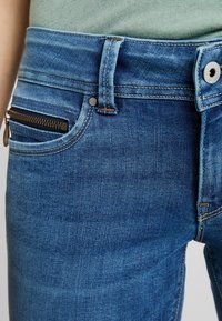 Jeans in denim blu scuro con una vestibilità slim, una tasca frontale con zip nera e cuciture a contrasto lungo la cintura e le cuciture.