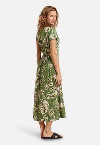 Groene maxi-jurk met bladranden, korte mouwen en een getailleerde taille. Soepele textuur, gecombineerd met zwarte platte sandalen.