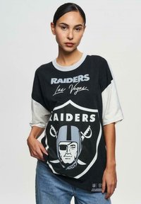Svart och vit överdimensionerad T-shirt med en Raiders-logotyp, texten "Las Vegas" och en grafik av en hjälmad figur på ett sköld.