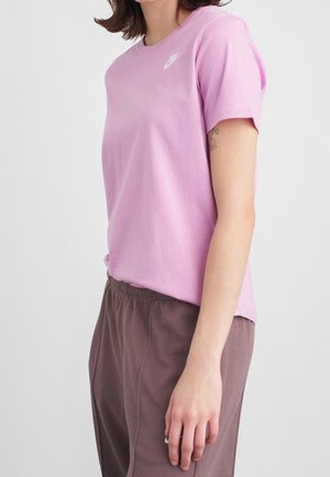 Personne portant un t-shirt Nike violet clair et un pantalon marron, debout les bras détendus le long du corps devant un fond uni.