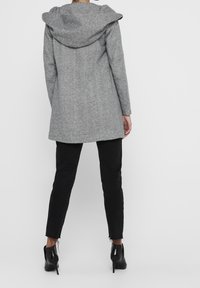 Grau Wollmantel mit großer Kapuze, taillierter Passform und leicht längerem Rückenteil, getragen über schwarzen Slim-Fit-Hosen und High-Heels.