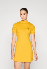Žlté šaty s krátkym rukávom a vysokým golierom, s fitted siluetou, na ktorých je malý čierny vyšitý smajlík na hrudi.