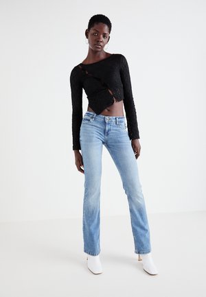 Must pikkade varrukatega lühike top diagonaalsete lõigetega, helesinised sirged teksapüksid ja valged kannaosaga pahkluu saapad, mida kannab seisvas asendis naine.