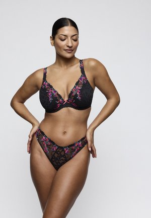 Woman posing in black lace lingerie with colorful floral embroidery, standing with hands on hips against plain light background.