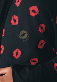 Sort stof mønstret med røde og ét guld læbestift kysaftryk, med en hånd, der hviler blidt på siden.