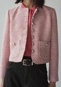 Lyserød tweedjakke med krave, sølvknapper, to forlommer og en tætsiddende pasform. Båret over en lyserød polo og mørk denim.