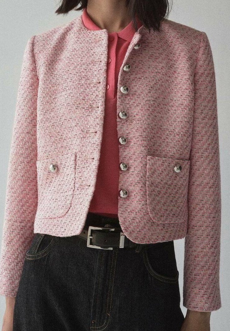 Lyserød tweedjakke med krave, sølvknapper, to forlommer og en tætsiddende pasform. Båret over en lyserød polo og mørk denim.