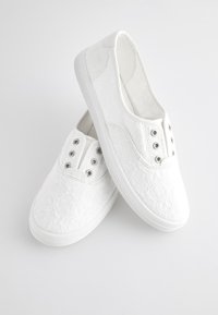 Paire de chaussures en toile blanches à enfiler avec motif en dentelle florale et œillets métalliques, placées sur fond blanc.