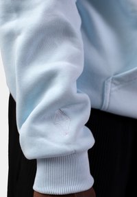 Ljusblå sweatshirtärm med ribbad mudd och en vit geometrisk logotyp broderad på underarmen. Slät tygtextur.