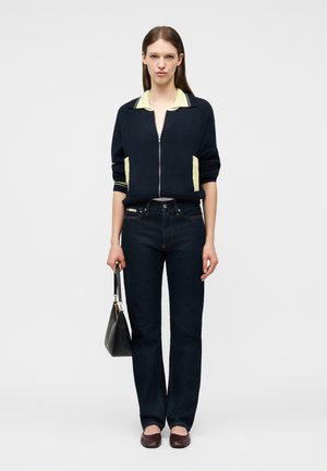 Mlada ženska stoji oblečena v temno jopico z zadrgo z svetlo rumenimi poudarki, temne kavbojke, rjave čevlje in drži črno torbico.