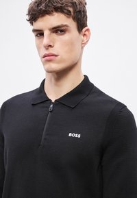Črna polo majica z zadrgo iz teksturirane tkanine, z ostrim ovratnikom in belim napisom "BOSS" na levi strani prsnega koša.