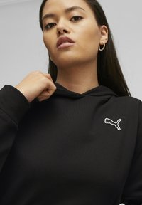 Felpa con cappuccio nera dal texture morbido, con il logo Puma bianco sul petto. Il modello sta afferrando il cappuccio, mettendo in mostra il design.