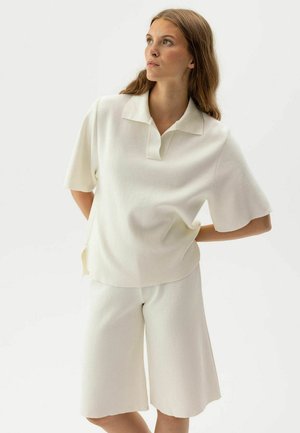 Sieviete ar gariem matiem, valkājot vaļīgu baltu īsroku krekliņu un tumši saskaņotas ceļa garuma šortus, stāv ar rokām aiz muguras, skatoties pa labi.