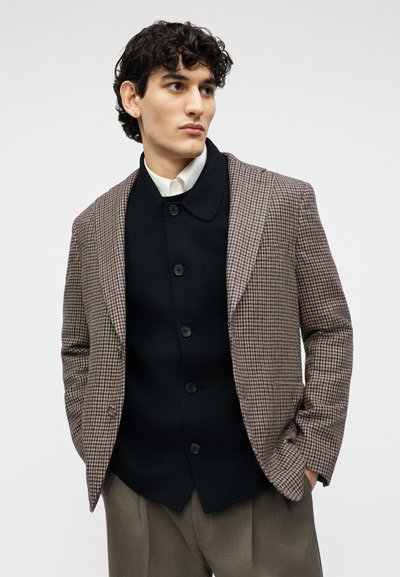 Νέος άνδρας με σγουρά σκούρα μαλλιά, φοράει καφέ καρό σακάκι, μαύρο κουμπωμένο μπουφάν, λευκό πουκάμισο και γκρι παντελόνι, με τα χέρια στις τσέπες.