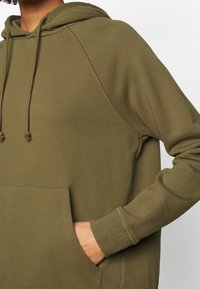 Olijfgroene capuchontrui met een kangoeroezak, geribde boorden en een trekkoord. Zacht materiaal met een gladde textuur en raglanmouwen.